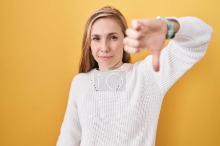 Foto de Young caucasian woman wearing white sweater over yellow background looking unhappy and angry showing rejection and negative with thumbs down gesture. bad expression. - Imagen libre de derechos