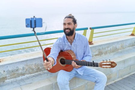 Foto de Young hispanic man musician holding classical guitar having video call at seaside - Imagen libre de derechos
