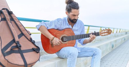 Foto de Young hispanic man musician playing classical guitar sitting on bench at seaside - Imagen libre de derechos