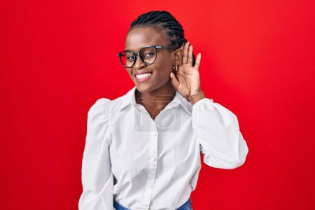 Téléchargez les photos : African woman with braids standing over red background smiling with hand over ear listening an hearing to rumor or gossip. deafness concept. - en image libre de droit