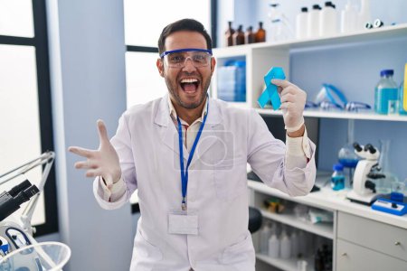 Téléchargez les photos : Jeune homme hispanique avec barbe travaillant au laboratoire scientifique tenant un ruban bleu célébrant la victoire avec un sourire heureux et l'expression gagnante avec les mains levées - en image libre de droit