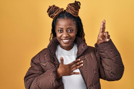 Téléchargez les photos : African woman with braided hair standing over yellow background smiling swearing with hand on chest and fingers up, making a loyalty promise oath - en image libre de droit