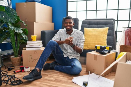 Téléchargez les photos : African american man sitting on the floor at new home with hands together and crossed fingers smiling relaxed and cheerful. success and optimistic - en image libre de droit