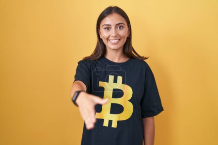 Foto de Mujer hispana joven con camiseta bitcoin sonriente amigable ofreciendo apretón de manos como saludo y bienvenida. negocios exitosos. - Imagen libre de derechos