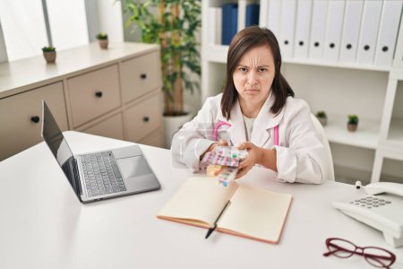 Photo for Hispanic girl with down syndrome wearing doctor uniform and stethoscope holding pills depressed and worry for distress, crying angry and afraid. sad expression. - Royalty Free Image