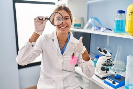 Photo for Young blonde woman working at scientist laboratory using magnifying glass smiling happy and positive, thumb up doing excellent and approval sign - Royalty Free Image