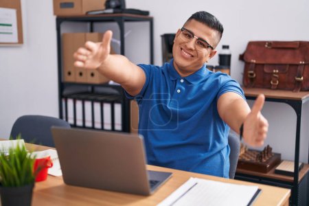 Téléchargez les photos : Young hispanic man working at the office with laptop looking at the camera smiling with open arms for hug. cheerful expression embracing happiness. - en image libre de droit