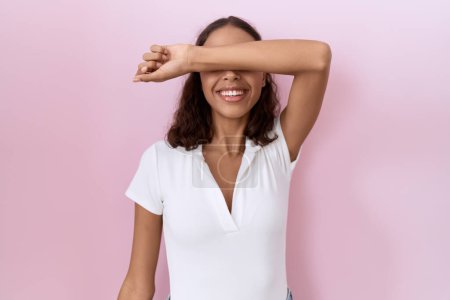 Foto de Mujer hispana joven con camiseta blanca casual cubriendo los ojos con el brazo sonriendo alegre y divertido. concepto ciego. - Imagen libre de derechos