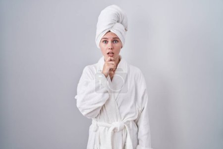 Photo for Blonde caucasian woman wearing bathrobe looking fascinated with disbelief, surprise and amazed expression with hands on chin - Royalty Free Image