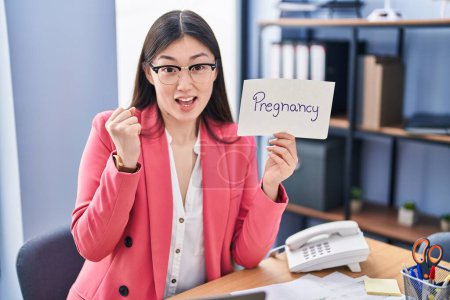 Photo for Chinese young woman working at the office holding pregnancy sign screaming proud, celebrating victory and success very excited with raised arms - Royalty Free Image
