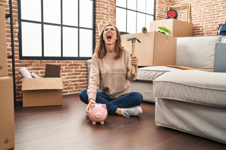 Photo for Young woman holding piggy bank and hammer moving to a new home angry and mad screaming frustrated and furious, shouting with anger looking up. - Royalty Free Image