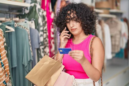 Téléchargez les photos : Young middle eastern woman customer talking on smartphone holding credit card at clothing store - en image libre de droit