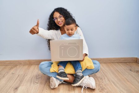 Foto de Young hispanic mother and kid using computer laptop sitting on the floor approving doing positive gesture with hand, thumbs up smiling and happy for success. winner gesture. - Imagen libre de derechos