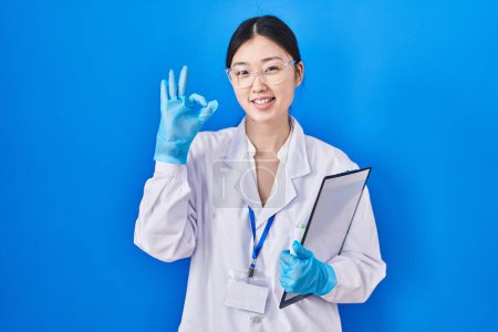 Photo for Chinese young woman working at scientist laboratory smiling positive doing ok sign with hand and fingers. successful expression. - Royalty Free Image