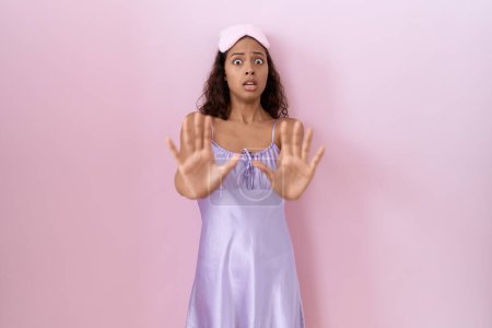 Young hispanic woman wearing sleep mask and nightgown afraid and terrified with fear expression stop gesture with hands, shouting in shock. panic concept. 