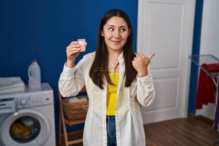 Téléchargez les photos : Jeune femme latine faisant la lessive tenant détergent pointant pouce vers le côté souriant heureux avec bouche ouverte - en image libre de droit