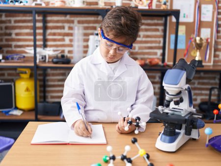 Foto de Adorable estudiante hispano escribiendo en un cuaderno sosteniendo moléculas en el aula de laboratorio - Imagen libre de derechos