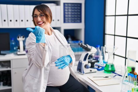 Téléchargez les photos : Femme enceinte travaillant au laboratoire scientifique souriant avec le visage heureux regardant et pointant vers le côté avec le pouce vers le haut. - en image libre de droit
