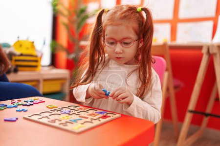 Foto de Adorable chica pelirroja jugando con matemáticas juego de puzzle sentado en la mesa en el jardín de infantes - Imagen libre de derechos