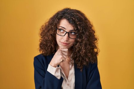 Foto de Hispanic woman with curly hair standing over yellow background with hand on chin thinking about question, pensive expression. smiling with thoughtful face. doubt concept. - Imagen libre de derechos