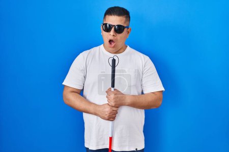 Téléchargez les photos : Hispanic young blind man holding cane in shock face, looking skeptical and sarcastic, surprised with open mouth - en image libre de droit