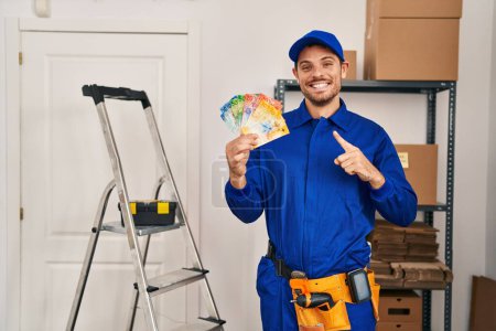 Téléchargez les photos : Young hispanic man working at renovation holding swiss banknotes smiling happy pointing with hand and finger to the side - en image libre de droit