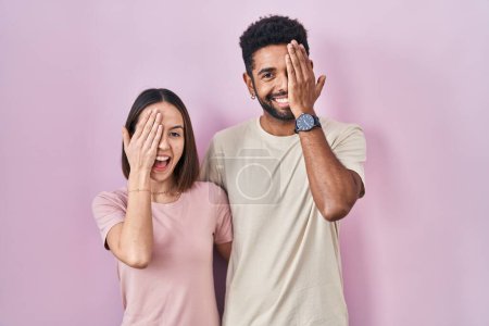 Foto de Joven pareja hispana junta sobre fondo rosa cubriendo un ojo con la mano, sonrisa confiada en la cara y emoción sorpresa. - Imagen libre de derechos