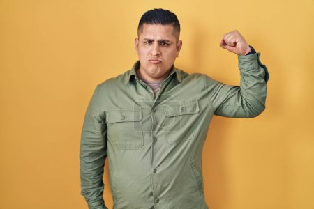 Photo for Hispanic young man standing over yellow background strong person showing arm muscle, confident and proud of power - Royalty Free Image