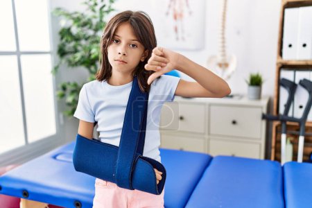Foto de Young hispanic girl wearing arm on sling at rehabilitation clinic with angry face, negative sign showing dislike with thumbs down, rejection concept - Imagen libre de derechos