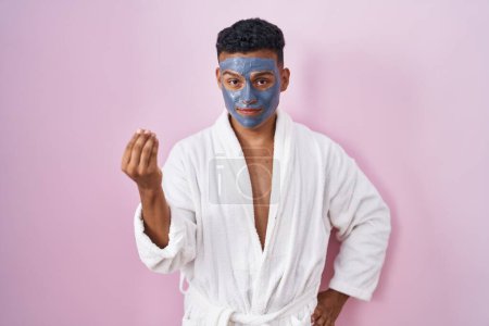 Téléchargez les photos : Jeune homme hispanique portant un masque de beauté et peignoir de bain faisant geste italien avec la main et les doigts expression confiante - en image libre de droit
