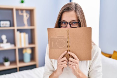 Photo for Young woman covering face with book sitting on bed at bedroom - Royalty Free Image