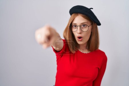 Téléchargez les photos : Jeune rousse femme debout portant des lunettes et un béret pointant du doigt surpris devant, bouche ouverte expression étonnée, quelque chose sur le devant - en image libre de droit