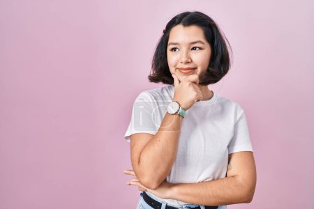 Photo for Young hispanic woman wearing casual white t shirt over pink background looking confident at the camera smiling with crossed arms and hand raised on chin. thinking positive. - Royalty Free Image