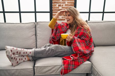 Foto de Mujer joven sentada en el sofá tomando un café en casa muy feliz y sonriente mirando lejos con la mano sobre la cabeza. concepto de búsqueda. - Imagen libre de derechos