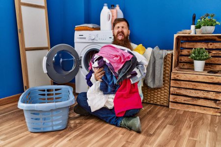Foto de Redhead man with long beard putting dirty laundry into washing machine sticking tongue out happy with funny expression. - Imagen libre de derechos