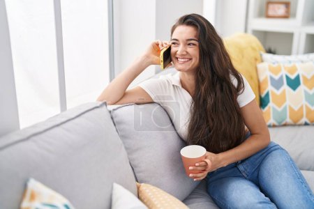 Téléchargez les photos : Jeune femme hispanique boire du café et parler sur le smartphone à la maison - en image libre de droit