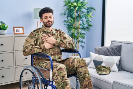 Téléchargez les photos : Homme arabe portant un uniforme de camouflage de l'armée assis sur un fauteuil roulant souriant avec les mains sur la poitrine avec les yeux fermés et geste reconnaissant sur le visage. concept de santé. - en image libre de droit