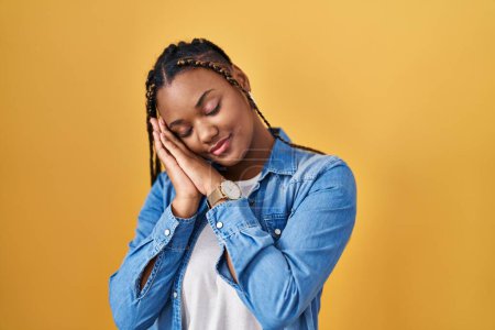 Téléchargez les photos : Femme afro-américaine avec des tresses debout sur fond jaune endormi fatigué rêvant et posant avec les mains ensemble tout en souriant avec les yeux fermés. - en image libre de droit
