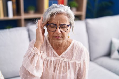 Foto de Mujer de pelo gris de mediana edad que sufre de dolor de cabeza sentada en el sofá en casa - Imagen libre de derechos
