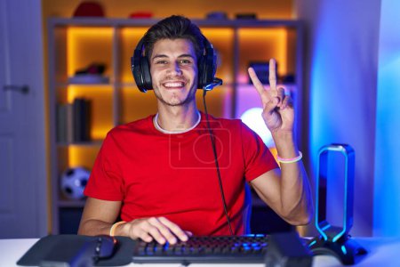 Foto de Young hispanic man playing video games showing and pointing up with fingers number two while smiling confident and happy. - Imagen libre de derechos
