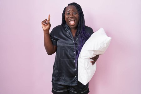 Photo for Young african woman wearing pijama hugging pillow smiling amazed and surprised and pointing up with fingers and raised arms. - Royalty Free Image