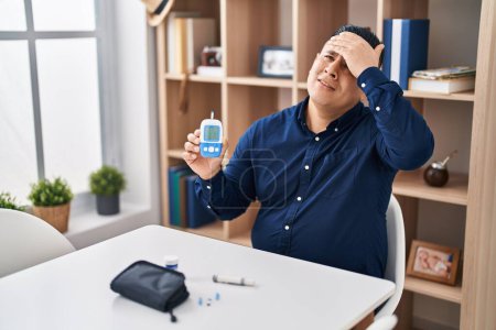 Téléchargez les photos : Hispanic young man holding glucometer device stressed and frustrated with hand on head, surprised and angry face - en image libre de droit