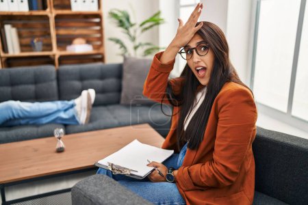 Foto de Young hispanic woman working as psychology counselor surprised with hand on head for mistake, remember error. forgot, bad memory concept. - Imagen libre de derechos