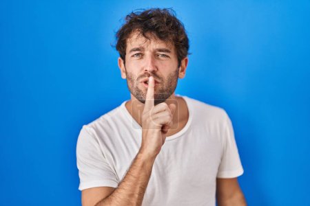 Photo for Hispanic young man standing over blue background asking to be quiet with finger on lips. silence and secret concept. - Royalty Free Image