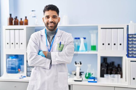 Téléchargez les photos : Jeune homme hispanique portant l'uniforme de scientifique debout avec les bras croisés geste au laboratoire - en image libre de droit
