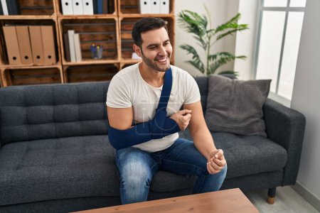 Foto de Young hispanic man with beard wearing arm on sling sitting at therapy consult looking positive and happy standing and smiling with a confident smile showing teeth - Imagen libre de derechos