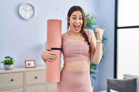 Photo for Young brunette woman holding yoga mat pointing thumb up to the side smiling happy with open mouth - Royalty Free Image