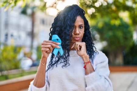 Photo for Middle age hispanic woman holding blue ribbon serious face thinking about question with hand on chin, thoughtful about confusing idea - Royalty Free Image