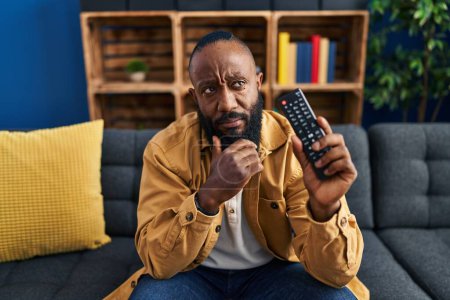 Photo for African american man holding television remote control serious face thinking about question with hand on chin, thoughtful about confusing idea - Royalty Free Image