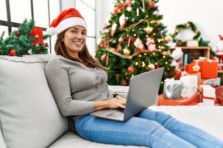Foto de Mujer latina joven usando portátil sentado junto al árbol de Navidad en casa - Imagen libre de derechos